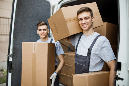 Two young handsome smiling workers wearing uniforms are standing in front of the van full of boxes holding boxes in their hands. House move, mover service.